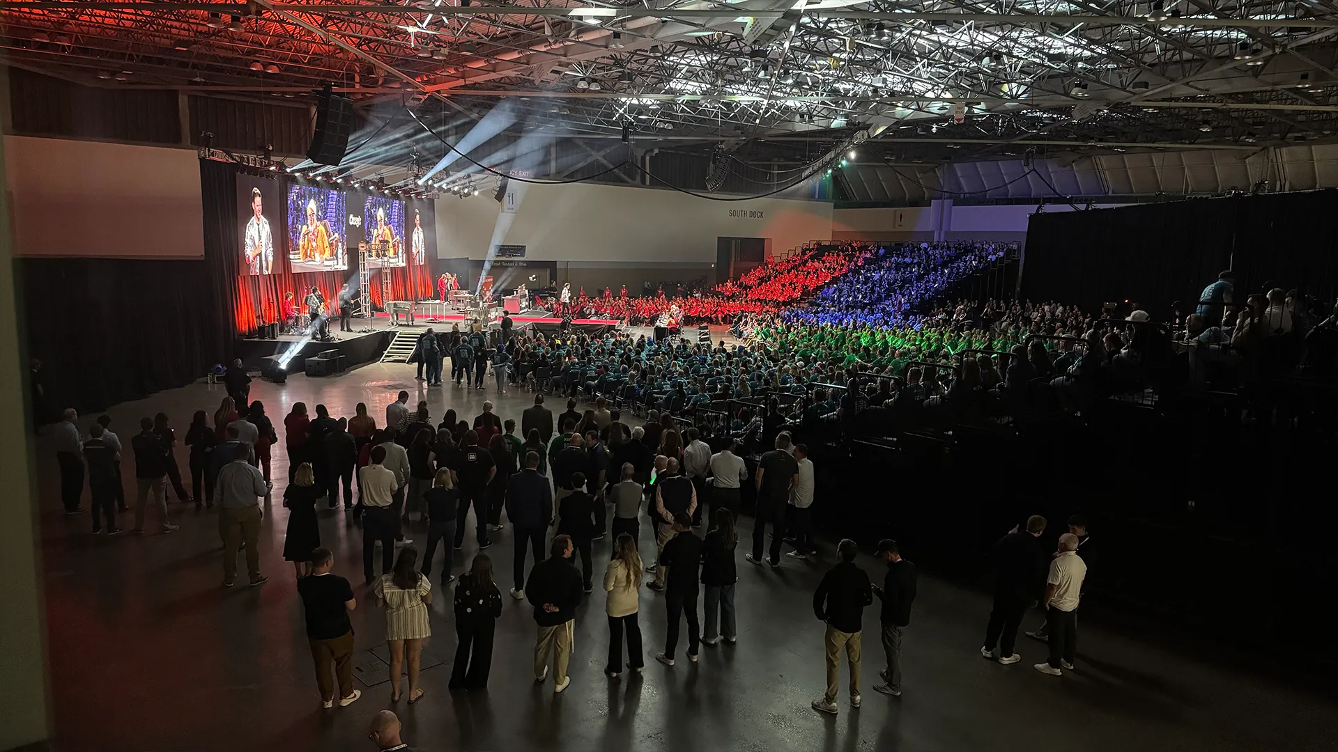 Large audience watching a corporate pizza competition with live video displays, stage lighting, and audio production