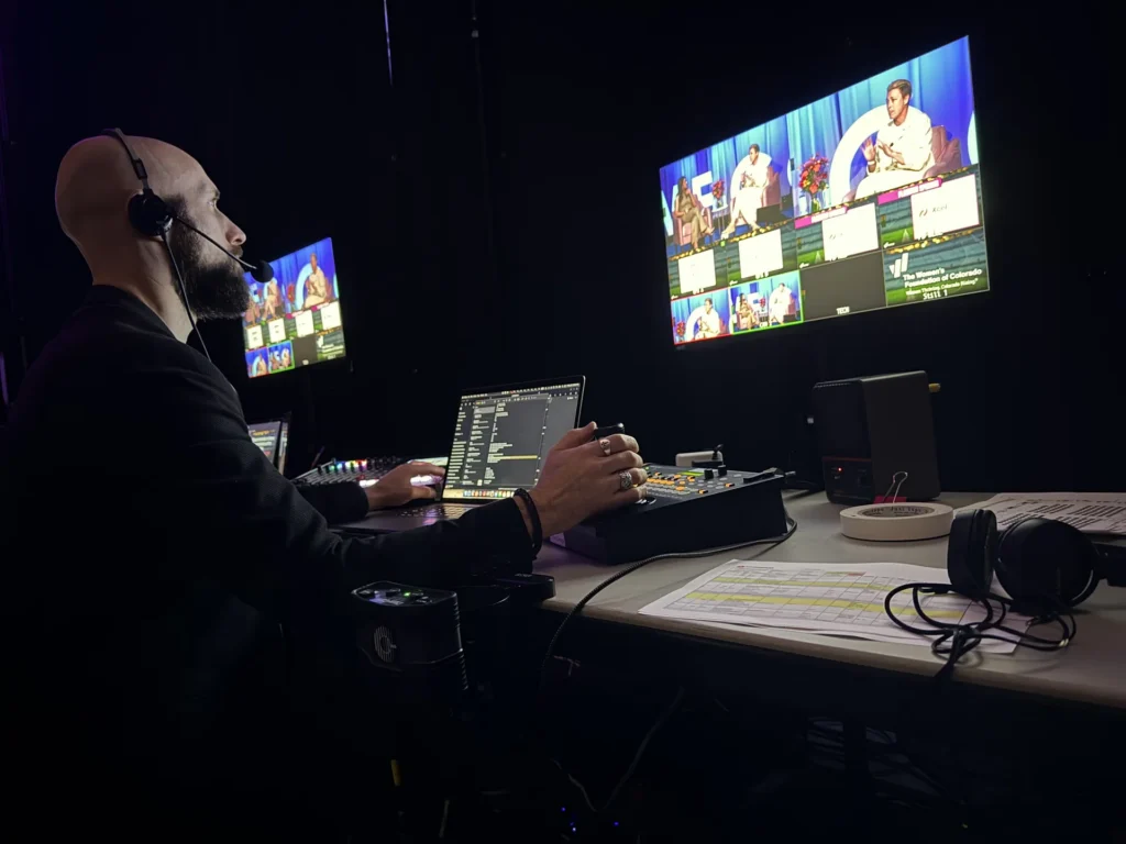 A man wearing a headset controls video switching equipment in a dark room with two monitors showing a panel discussion.